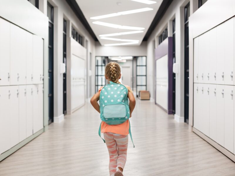 Rear,View,Of,Elementary,Schoolgirl,With,Backpack,Walking,Amidst,Lockers